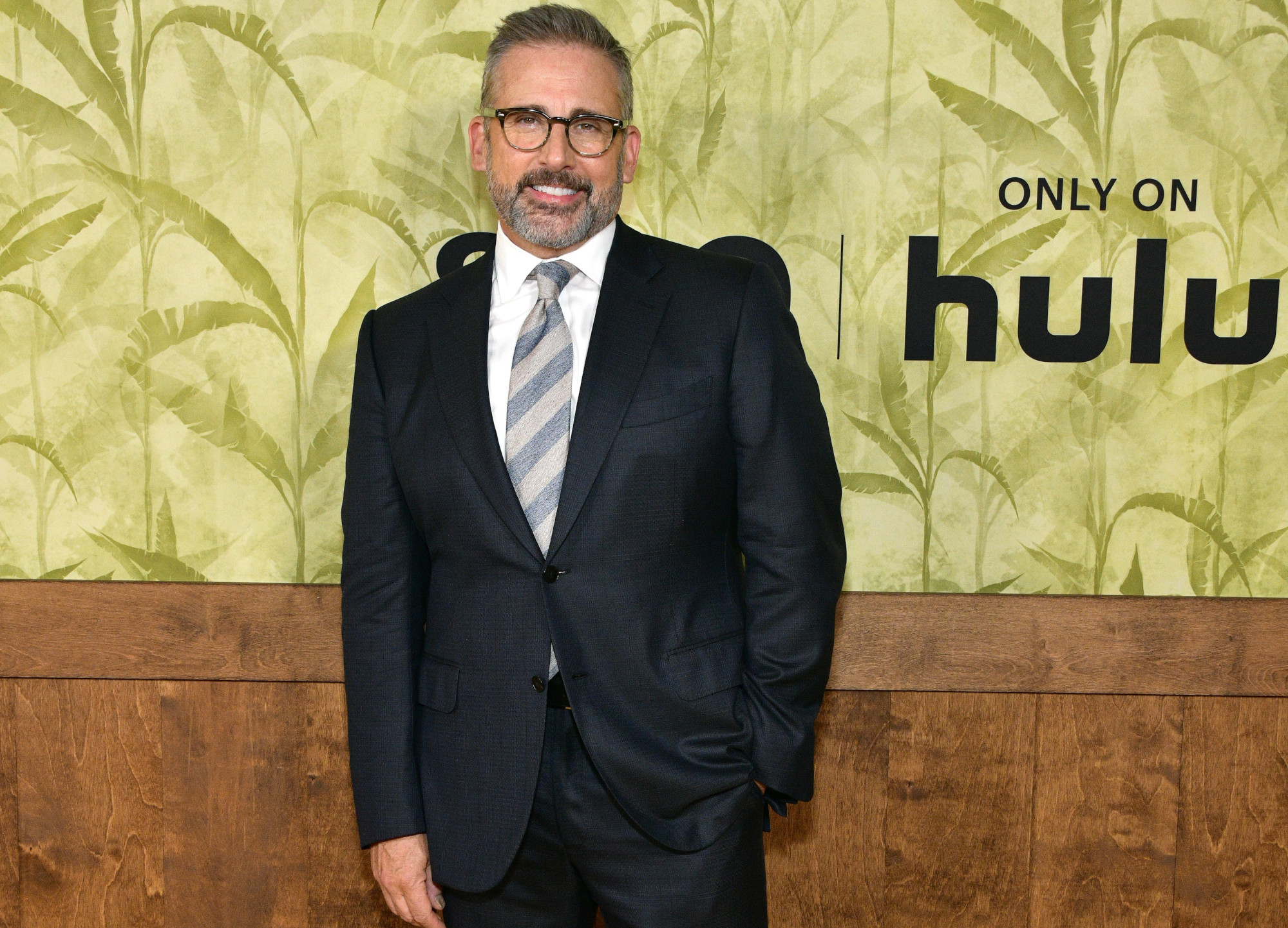 史蒂夫·卡瑞尔在《病人》首映式上。他穿着黑色西装，系着灰色条纹领带，站在一面金棕色相间的墙上，上面写着“Hulu”。
