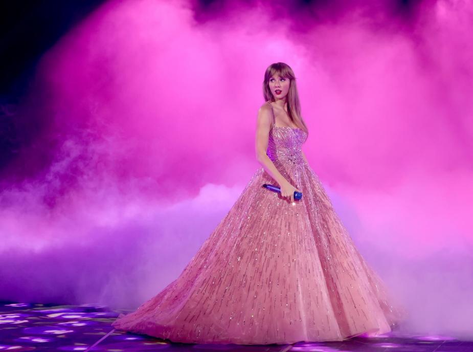 泰勒·斯威夫特在“时代之旅”中身穿粉色舞会礼服站在舞台上，周围是粉红色的薄雾。